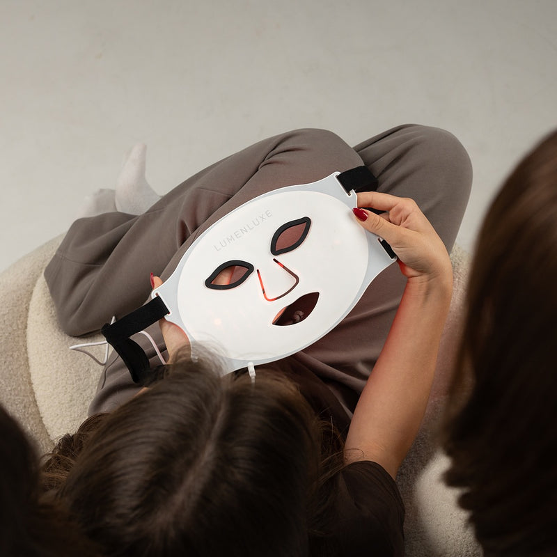Žena drží LumenLuxe LED masku na tvári pri relaxácii doma — červené svetlo terapia pleti
